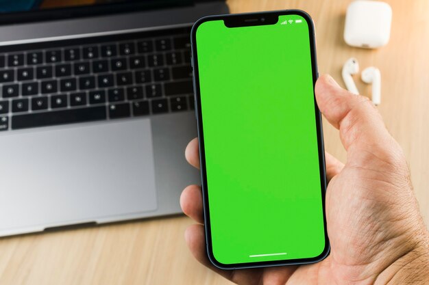 Main masculine tenant un smartphone avec un écran vert sur fond en bois avec un ordinateur. Vue de dessus. Clé chroma.