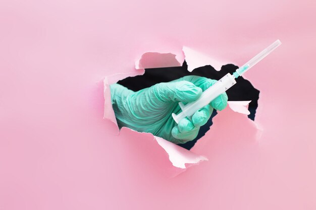 la main d'un médecin tient une seringue avec un vaccin à travers un trou dans du papier déchiré sur fond rose