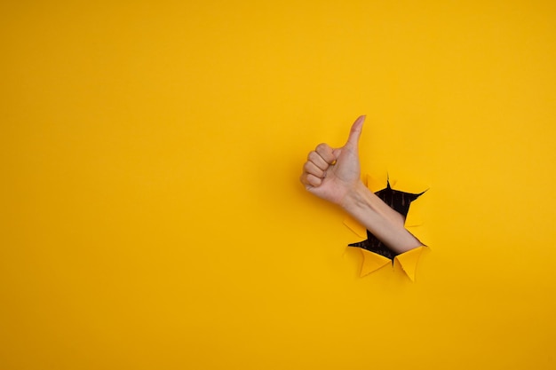 Main montrant un pouce vers le haut à travers un trou déchiré dans un mur de papier jaune. Bravo, bon concept de travail.