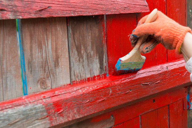 Une main de peintre peignant une clôture en bois de couleur rouge