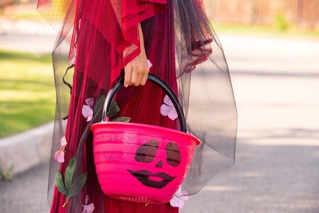 Main de petite fille en costume de sorcière d'halloween tenant un panier en plastique rose ou un seau plein de friandises en se tenant debout sur la route