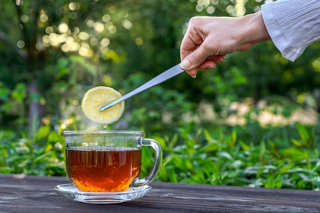 Photo la main avec des pinces met un citron dans le thé dans une tasse en verre sur une table en bois