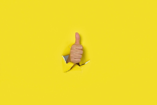 La main avec le pouce en l'air en signe d'approbation sortant du trou d'un fond de papier jaune déchiré