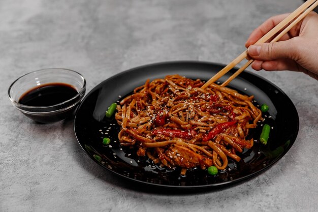 Main prenant udon avec du poulet allongé sur une assiette noire et de la sauce soja dans un bol en verre.