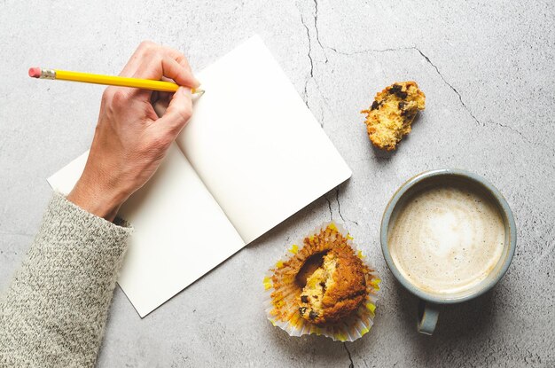 Une main qui écrit sur un cahier avec un crayon, une tasse de latte et un muffin, sur fond gris lapidé.