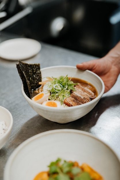 Main de serveur méconnaissable servant des ramen de soupe de nouilles asiatiques avec de la viande de tofu légumes nori et oeuf
