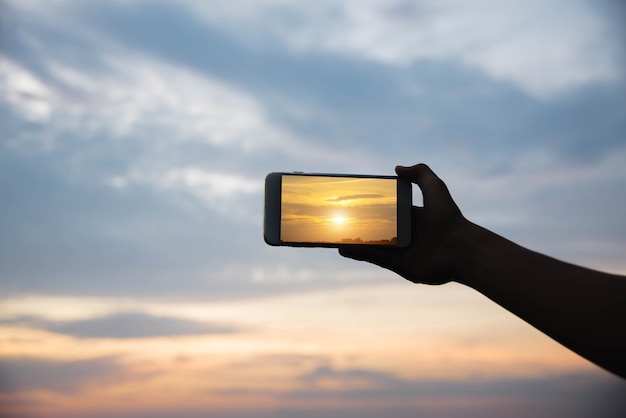 Main de silhouette tenant le smartphone prendre une photo au coucher du soleil.