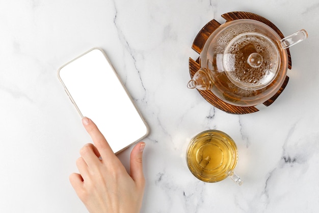 La main tape sur le téléphone près d'une tasse en verre avec une tisane verte et une théière sur fond de marbre blanc. Vue de dessus. Discuter au téléphone