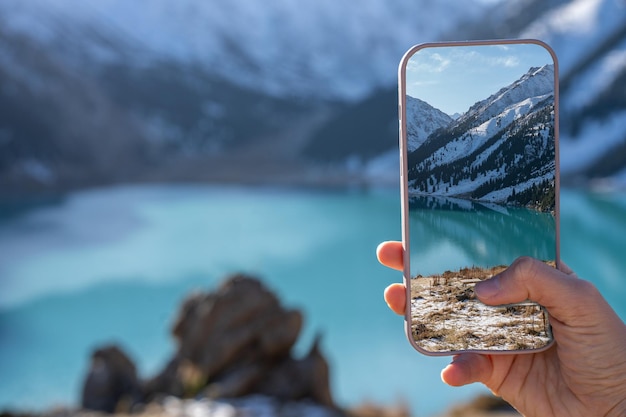 Main avec un téléphone prenant l'image d'un lac turquoise entouré de montagnes