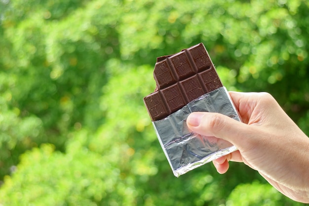 Main Tenant Une Barre De Chocolat Noir Dans Un Paquet D'aluminium Avec Un Feuillage Vert Flou En Toile De Fond