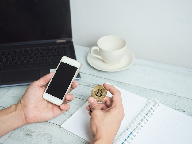 Main tenant un bitcoin d'or et un téléphone portable avec un espace de travail sur fond de table en bois blanc