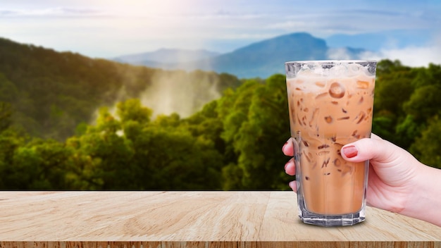 Main tenant le café glacé en verre sur une table en bois avec fond de nature de paysage Café au lait glacé