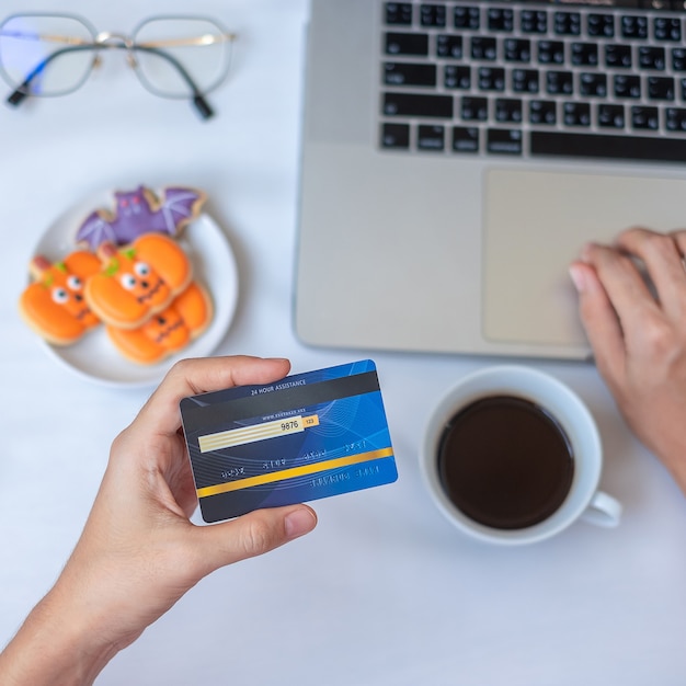 Main tenant une carte de crédit pour faire des achats en ligne sur un ordinateur portable pendant que vous mangez des biscuits d'Halloween et du café. Joyeux Halloween, bonjour octobre, automne automne, concept festif, fête et vacances