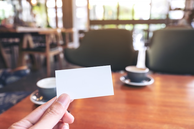 Photo une main tenant une carte de visite vide blanche avec deux tasses à café sur une table en bois au café