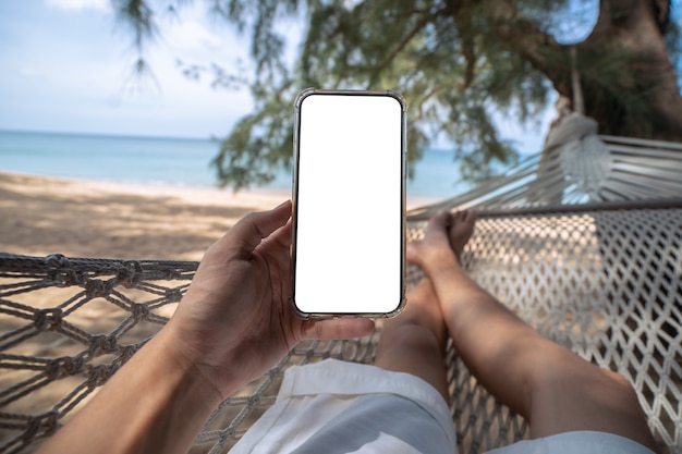 Main tenant une maquette mobile avec un écran blanc tout en s'étendant sur une balançoire hamac entre les arbres sur la plage.