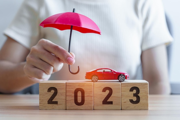 Main tenant un parapluie et une couverture jouet de voiture rouge avec un bloc de l'année 2023 sur la table Réparation de garantie d'assurance automobile Argent bancaire financier et concept du Nouvel An