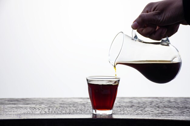 Photo la main tenant un pot de café noir versé dans un verre sur une table en bois