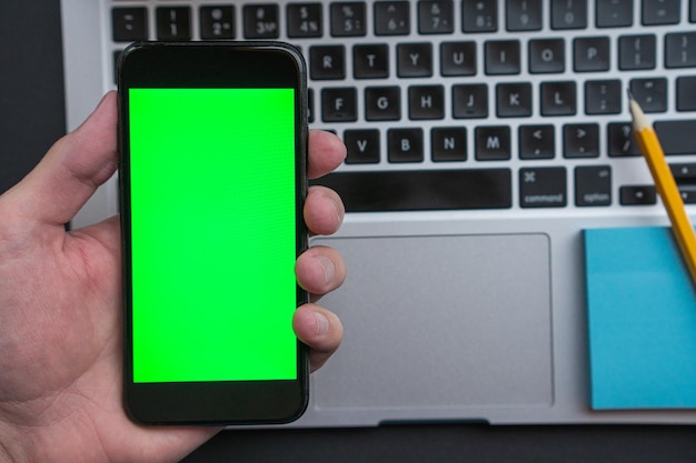 Main tenant un Smartphone avec affichage de maquette d'écran vert. En arrière-plan se trouve un ordinateur portable