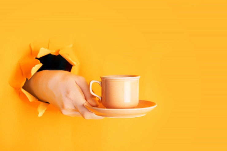 Photo main tenant une tasse de café à travers un trou dans un mur déchiré en safran ou orange clair
