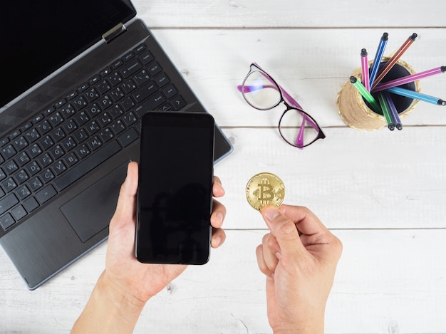 Main tenant un téléphone portable et un bitcoin d'or avec des lunettes pour ordinateur portable et une vue de dessus de fond de boîte à stylos