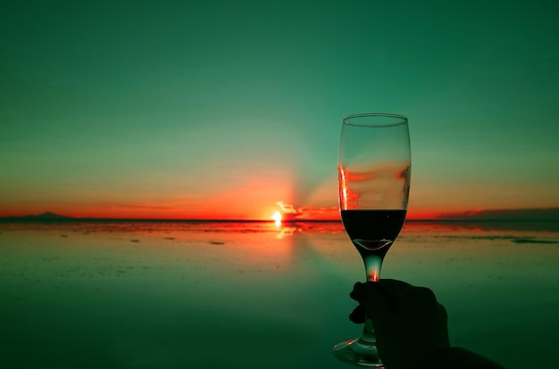 Main tenant un verre à vin contre le coucher du soleil sur l'effet miroir des salines d'Uyuni en Bolivie