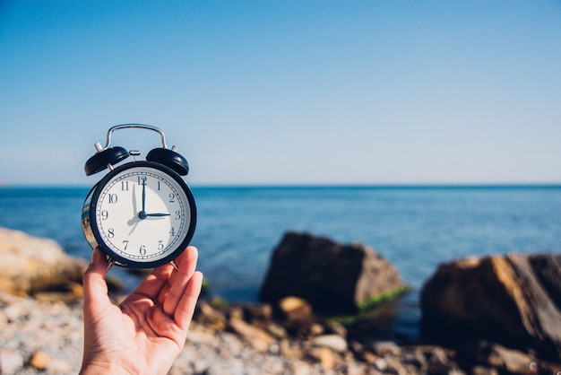 Main tenir la montre sur fond de plage. Réveil sur fond de mer et de sable à différentes heures avec les vacances d'été. Concept de temps de vacances.