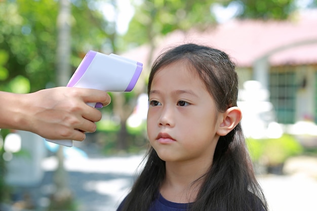 Main avec thermomètre infrarouge mesurant le front d'une petite fille asiatique avant d'entrer dans l'aire de jeux