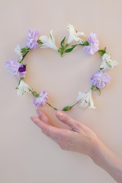 La main tient le cadre du coeur fait de clous de girofle et d'alstroemeria, de branches, de feuilles et de pétales sur fond beige. Mise à plat, vue de dessus