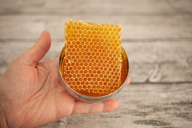 Une main tient du miel dans des nids d'abeilles dans un bol doré sur un fond en bois.