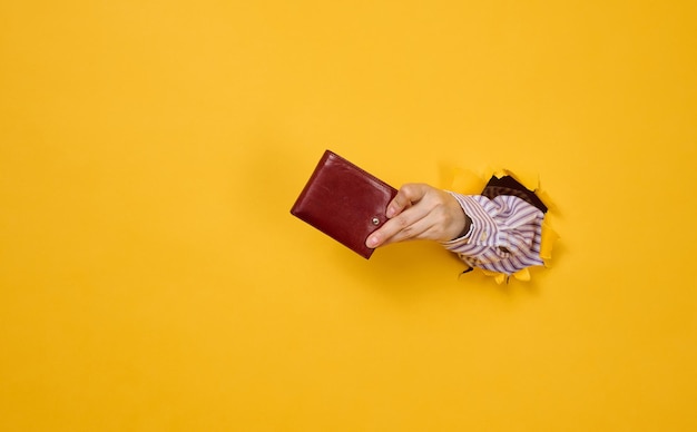 La main tient un portefeuille en cuir marron sur fond jaune, une partie du corps sort d'un trou déchiré dans le papier