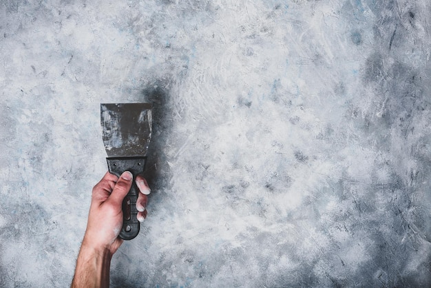 La main tient une spatule sale. Fond de mur de béton gris.