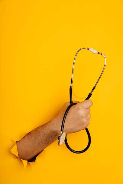 La main tient un stéthoscope hors d'un trou déchiré dans l'image verticale du mur de papier jaune