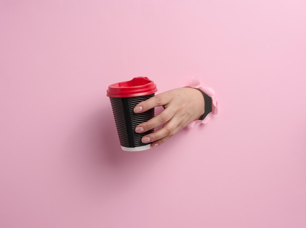 La main tient la tasse brune en carton de papier pour le café, fond rose. Vaisselle écologique, zéro déchet, partie du corps sortant d'un trou déchiré dans le papier