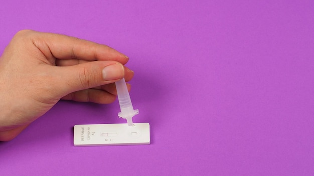 Une main tient un tube d'extraction d'antigène et un dispositif de test d'antigène Écouvillon stérilisé sur le sol avec un fond violetx9
