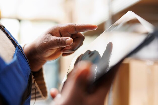 Main de travailleur d'entrepôt afro-américain tapant sur l'écran de la tablette numérique dans la salle de stockage. Bras de responsable de la logistique et de la distribution supervisant l'inventaire des marchandises à l'aide d'une application logicielle en gros plan