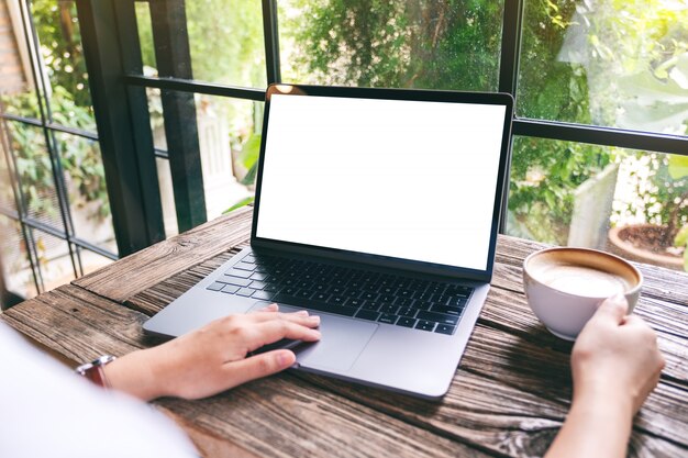 Une main en utilisant et en touchant le pavé tactile de l'ordinateur portable avec un écran de bureau blanc vierge tout en buvant du café