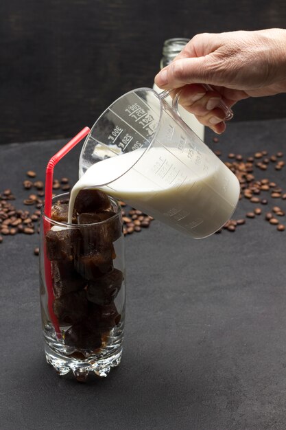 Photo main verse le lait d'une tasse à mesurer dans un verre avec des glaçons au café avec de la paille rouge.