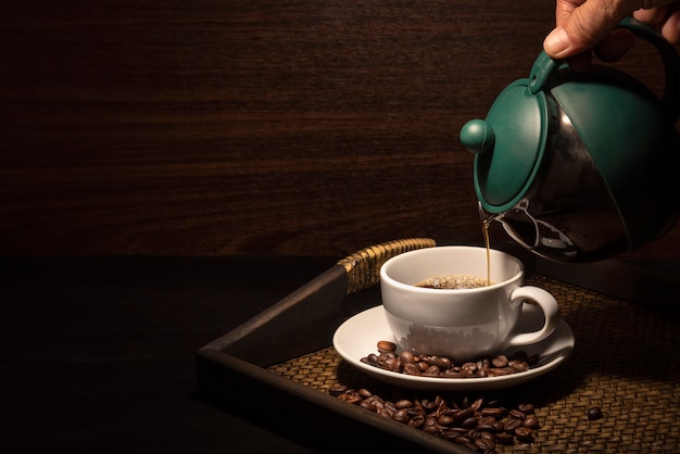 Main verser le café sur la tasse de café blanc avec des grains de café sur le plateau en bambou