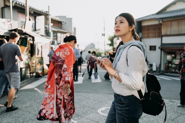 main de voyageur fille tenant un téléphone portable regardant le panneau routier pour trouver la direction de l'hôtel par carte en ligne. belle dame japonaise locale marchant en arrière-plan portant une robe kimono à fleurs rouge traditionnelle