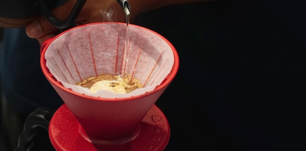 Mains de barista préparant un expresso chaud goutte à goutte en versant de l'eau chaude dans une tasse rouge avec du papier filtre goutte à goutte du café de la bouilloire sur fond noir TableBlack Espace pour le texte gros plan