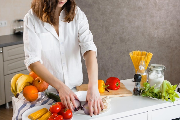 Les mains d'une belle jeune femme en chemise blanche dans la cuisine coupent des légumes poivrons rouges et jaunes maïs oignons bleus Végétariens aliments sains