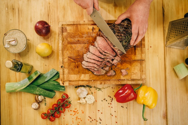 Mains de chef masculin avec couteau coupe la viande rôtie sur des tranches, vue de dessus.