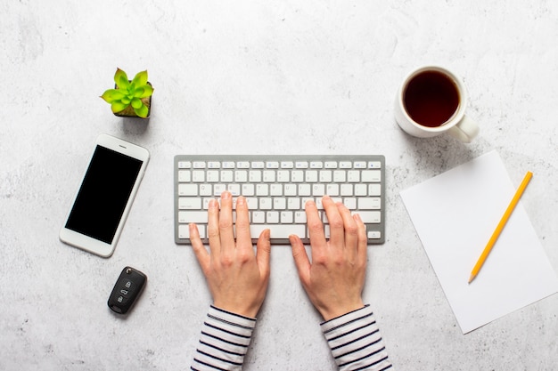 Mains sur le clavier, les clés de voiture, une tasse de café, une feuille de papier vierge, un crayon et une fleur de chambre sur un fond en béton.