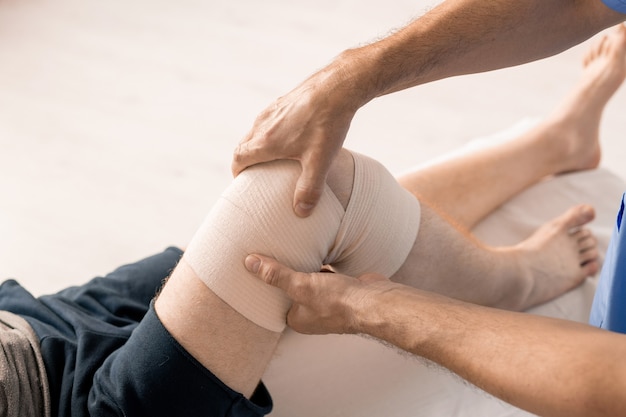 Mains d'un clinicien enveloppant le genou d'un patient handicapé avec un bandage flexible pendant la procédure médicale avant l'entraînement physique de rééducation