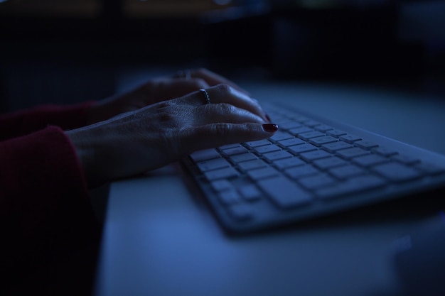 Des mains coupées d'une femme utilisant un clavier d'ordinateur sur une table dans une chambre noire à la maison