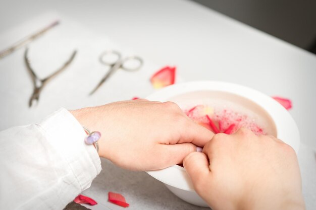 Les mains dans un bol blanc baignent les ongles avec de l'eau et des pétales de rose pour adoucir la cuticule et la peau sèche