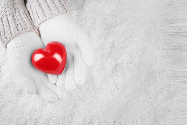 Mains dans des gants blancs chauds tenant un coeur rouge sur la neige