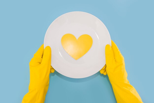 Des mains dans des gants jaunes tiennent une assiette blanche avec un coeur jaune isolé sur fond bleu. Vue d'en-haut.