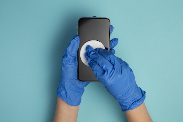 Mains dans des gants médicaux essuyez l'écran du smartphone avec un chiffon désinfectant Gros plan des mains nettoient le téléphone portable isolé sur fond bleu Désinfection du smartphone