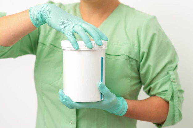 Les mains dans les gants de protection de l'esthéticienne ouvrent un pot de crème pour le corps blanc sur fond blanc.